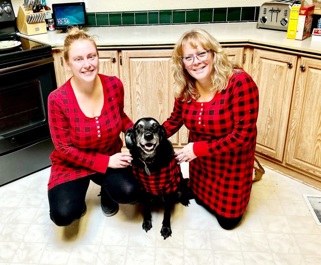 Two women and a dog. All three of them are wearing red flannel pajamas. The dog is a black lab and is smiling looking at the camera.